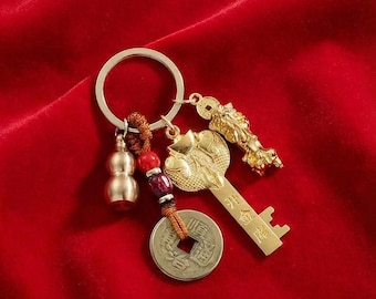 May include: A gold-colored keychain with multiple charms, including a key, a coin, and a gourd-shaped pendant. The key has Chinese characters. The charms are attached to a silver-colored ring. The background is a red fabric.