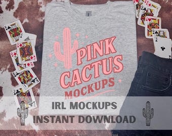 May include: A grey t-shirt featuring a pink cactus design and the text "Pink Cactus Mockups." Playing cards and dark denim jeans are arranged around the shirt. The words "IRL Mockups" and "Instant Download" are also visible.