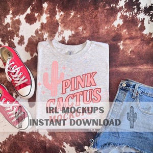 Puede incluir: Una camiseta gris claro con un gráfico de cactus rosa y las palabras "Pink Cactus". La imagen también incluye zapatillas rojas, pantalones cortos de mezclilla con un gráfico de cactus y el texto "IRL MOCKUPS INSTANT DOWNLOAD".