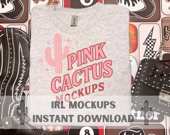May include: A grey t-shirt featuring a pink cactus design and the text "PINK CACTUS MOCKUPS IRL MOCKUPS INSTANT DOWNLOAD". The shirt is displayed with a black leather boot, a black purse, and a brown patterned background.