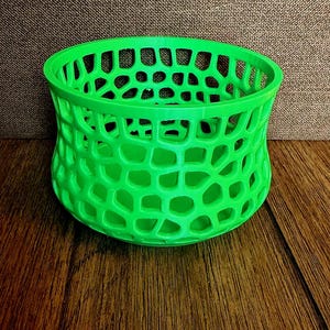 May include: A bright green, open-weave basket with a honeycomb-like pattern. The basket is cylindrical with a slightly flared top and sits on a wooden surface. The design allows for visibility of the contents.
