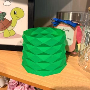 Op de afbeelding: Een felgroene, geometrisch gevormde plantenbak. De cilindrische container heeft een herhalend ruitpatroon. De plantenbak is gemaakt van een stevig materiaal en staat op een houten plank. De plantenbak is leeg en klaar voor een kleine plant.