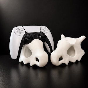 May include: A white and black PlayStation 5 controller sits above two white, bone-shaped decorative objects. The objects have a skull-like appearance, with eye sockets and a curved shape. The background is solid black.