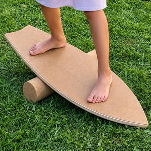Könnte beinhalten: Ein hellbraunes hölzernes Balance-Board mit einer glatten, flachen, surfbrettförmigen Oberfläche und einer separaten zylindrischen Rolle darunter. Das Board hat eine geschichtete Sperrholzkante und ist horizontal auf einer grünen Grasfläche platziert.