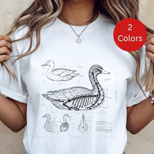 May include: White t-shirt featuring a detailed blueprint-style illustration of a duck, including its skeletal structure. The design is in black and white, with additional duck diagrams. A red circle with the text "2 Colors" is visible.