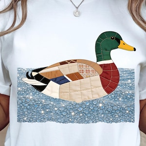 May include: White t-shirt featuring a colorful patchwork duck design floating on water. The duck has a green head, a yellow beak, and a body composed of various fabric patterns. The water is a light blue with sparkling details.