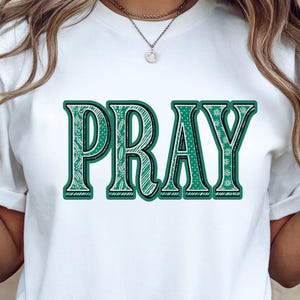 Pode incluir: Camiseta branca com a palavra "PRAY" em letras grandes, verdes e decorativas. A palavra é delineada em verde escuro e tem um design detalhado com padrões dentro das letras. A camiseta está sendo usada.