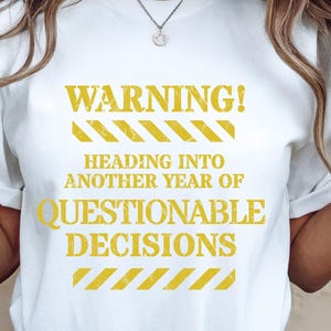 Op de afbeelding: Wit t-shirt met de gele tekst "WARNING! HEADING INTO ANOTHER YEAR OF QUESTIONABLE DECISIONS". De tekst is vetgedrukt met gele gevarenstrepen.