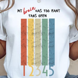 May include: White t-shirt with the text "MY brain HAS TOO MANY TABS OPEN" in black and red. Below the text are five vertical stripes in red, orange, yellow, green, and blue. The numbers 12345 are below the stripes.