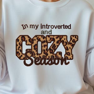 May include: White sweatshirt with the text "In my introverted and COZY Season" in brown and leopard print. The sweatshirt has a crew neck and long sleeves. A white mug is held in the left hand.