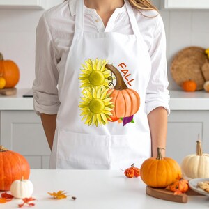 May include: White apron with a fall design. The design features two yellow sunflowers, an orange pumpkin, and the word "FALL" in brown. The apron has a pocket and is worn by a person.