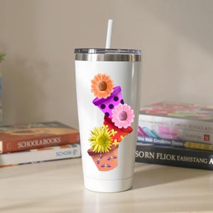 Peut inclure: Gobelet isotherme blanc avec un motif floral. Le motif présente des pots de fleurs empilés en violet, rose et rouge, chacun avec une fleur différente. Le gobelet a un couvercle argenté et une paille blanche.