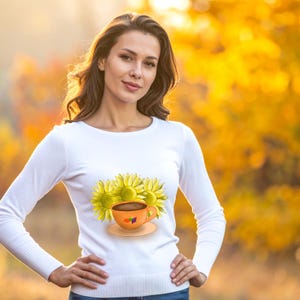May include: A white long-sleeved shirt with a graphic of three yellow sunflowers and an orange coffee cup on a saucer. The shirt is worn by a person with brown hair, against a blurred yellow and orange background.