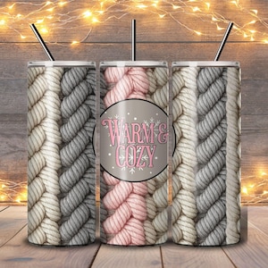 May include: Three stainless steel insulated tumblers with black straws. The tumblers feature a repeating pattern of thick, woven yarn in shades of gray, cream, and pink. The center tumbler has a circular design with the words "Warm & Cozy".