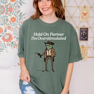 May include: A sage green t-shirt with the text "Hold On Partner I'm Overstimulated" and a cartoon frog wearing a cowboy hat and holding a gun. The frog is dressed in a brown jacket and belt.