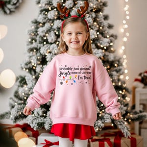 A young girl wearing a pink sweatshirt with a humorous Christmas-themed design stands in front of a decorated Christmas tree.