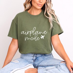 A woman wearing a green t-shirt with the text "airplane mode" and blue jeans, sitting on the floor with her legs crossed.