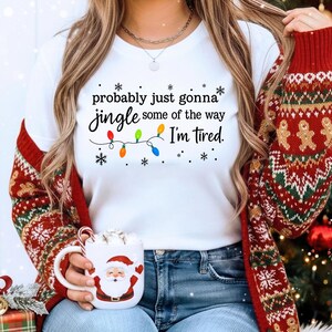 A woman wearing a white t-shirt with a humorous Christmas-themed text and a red and green sweater, holding a mug with a Santa Claus design.