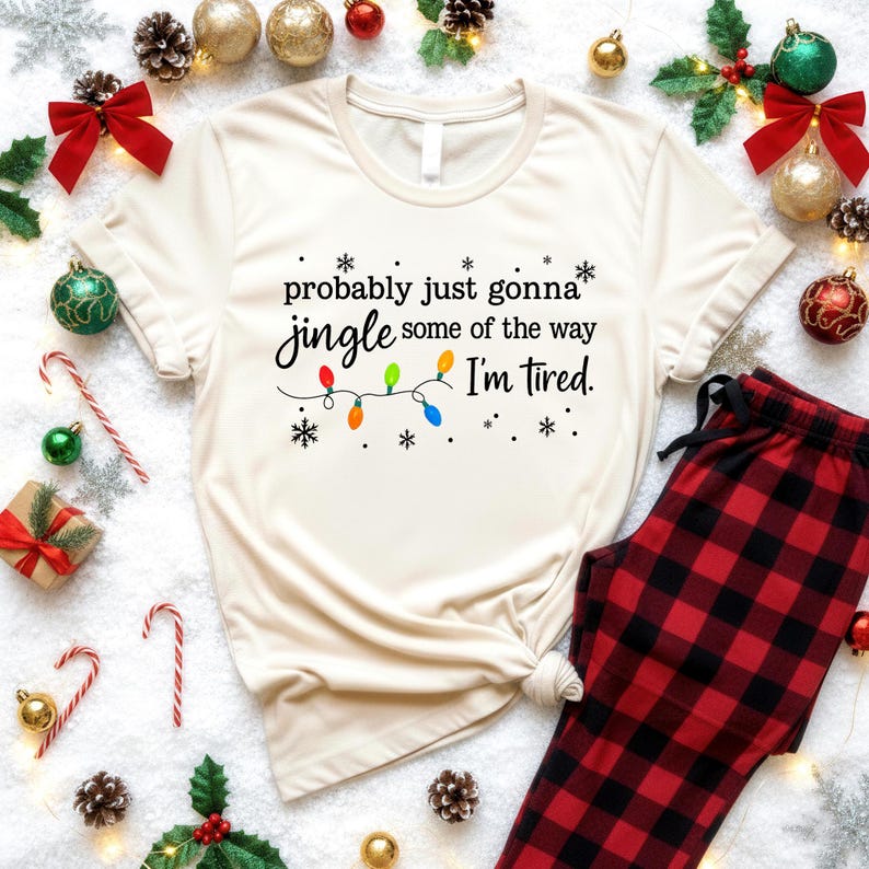 A white t-shirt with a humorous Christmas-themed text printed on it, surrounded by various Christmas decorations such as ornaments, pine cones, and a string of colorful lights.