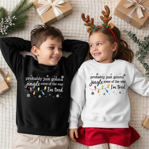 A young boy and girl are lying on a bed, surrounded by Christmas decorations and gifts. The boy is wearing a black sweatshirt with a humorous text, while the girl is wearing a white sweatshirt with a similar text.