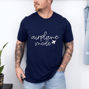 A man wearing a navy blue t-shirt with the text "airplane mode" printed on it, standing in front of a white wall.