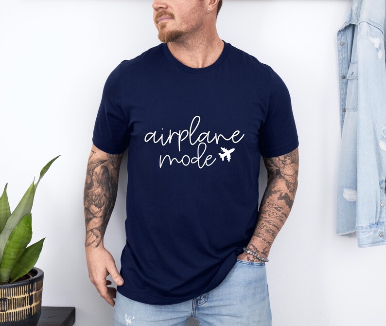 A man wearing a navy blue t-shirt with the text "airplane mode" printed on it, standing in front of a white wall.