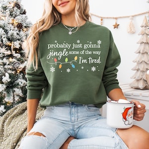 A woman wearing a green sweatshirt with a humorous Christmas-themed text printed on it, sitting in front of a Christmas tree.