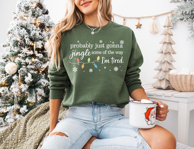 A woman wearing a green sweatshirt with a humorous Christmas-themed text printed on it, sitting in front of a Christmas tree.