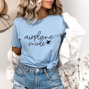 A woman wearing a light blue t-shirt with the text "airplane mode" printed on it, paired with denim shorts and a white cardigan. She is standing in front of a floral arrangement, with a straw hat visible in the background.