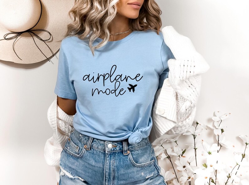 A woman wearing a light blue t-shirt with the text "airplane mode" printed on it, paired with denim shorts and a white cardigan. She is standing in front of a floral arrangement, with a straw hat visible in the background.