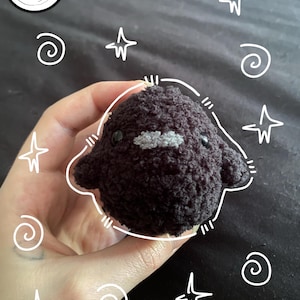 May include: A small, round, dark brown crocheted plush toy with a fuzzy texture, featuring two black plastic eyes and a small, light gray felt mustache detail. The plush is outlined with a white hand-drawn effect, surrounded by white stars and swirls.