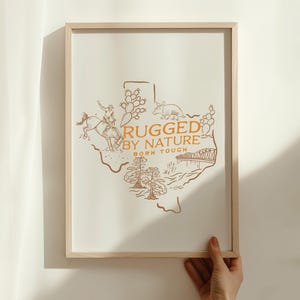 May include: A framed print featuring a detailed illustration of the state of Texas in a warm brown hue. The design includes a cowboy on a horse, an armadillo, and the text "RUGGED BY NATURE BORN TOUGH". The artwork is displayed in a light wooden frame.