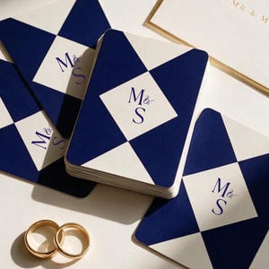 May include: A stack of rectangular cards and loose cards featuring a navy blue and cream diamond pattern with the monogram M&S in the centre. An envelope with the gold text MR & MRS and two gold metal rings are positioned next to the cards.