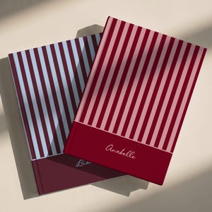 Könnte beinhalten: Zwei rechteckige Notizbücher mit vertikalen Streifen. Das obere Notizbuch ist rot und rosa mit dem in Weiß gedruckten Namen Anabelle. Das untere Notizbuch ist blau und weiß mit dem teilweise sichtbaren Namen Bia in Weiß auf dunkelrotem Grund.