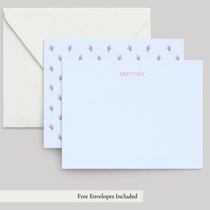 May include: A light blue stationery card featuring a repeating pattern of small pink flowers with green stems along the top and left edges, with the name BRITTNEY printed in pink in the center, accompanied by a plain white envelope.