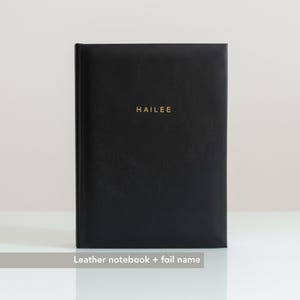 May include: A black leather notebook with the name "HAILEE" in gold foil lettering. The notebook is closed and has a textured surface. The notebook is on a white surface.
