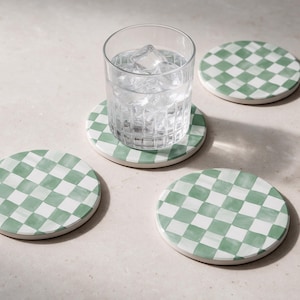 Op de afbeelding: Drie ronde onderzetters met een saliegroen en wit geblokt patroon, waarbij een helder glazen beker gevuld met ijsblokjes op een van de onderzetters staat. De onderzetters hebben een glad, plat oppervlak en een consistent rasterontwerp.