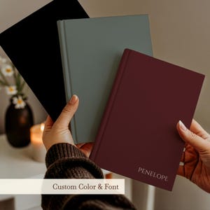 May include: Three hardcover journals in black, sage green, and deep burgundy. The burgundy journal features the name PENELOPE embossed in white text on the front cover. Each journal has a smooth, solid-colored finish and a vertical spine design.