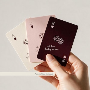 May include: Three playing cards in cream, light pink, and dark maroon. Each card features a heart suit symbol, a pair of dice with heart patterns, and the text oh how lucky we are. The cards are arranged in a fan shape with letters A, J, and E.