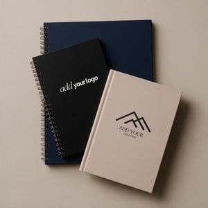 May include: A stack of three notebooks: a large dark blue spiral-bound notebook, a medium black spiral-bound notebook with the text "add your logo" in white, and a beige hardbound notebook with a black mountain logo and "ADD YOUR LOGO HERE".