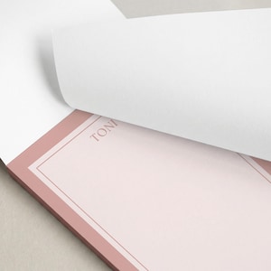 May include: A pink notepad with a double border and the initials "R.S." printed at the top. The notepad is rectangular and sits on a white surface.