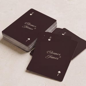 May include: A deck of dark brown playing cards with white text reading Eleanor & James. Each card features a white heart symbol at the top with the letter E and a white spade symbol at the bottom with the letter J. The cards have a smooth finish.