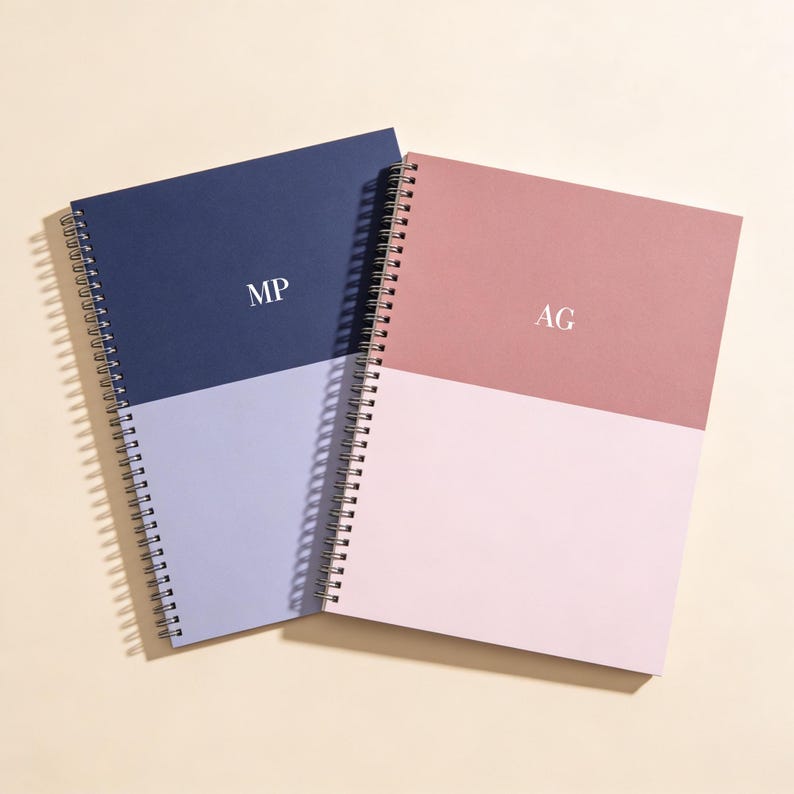 A personalized two tone burgundy and blush pink hardcover notebook with centered white monogram initials rests on layered cream fabrics beside a textured ceramic vase and a yellow billy ball flower. The colorblock design offers a warm feminine aesthetic ideal for journaling planners gifts and study notes. This premium minimalist stationery piece is photographed in soft natural lighting for an elegant cozy lifestyle look that appeals to modern desk decor lovers.