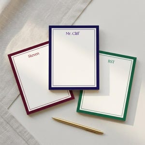 May include: A white notepad with a burgundy border and the initials "M.P." printed in the upper center. The notepad has a clean, minimalist design, suitable for writing notes or lists.