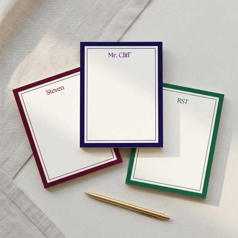 May include: A white notepad with a burgundy border and the initials "M.P." printed in the upper center. The notepad has a clean, minimalist design, suitable for writing notes or lists.