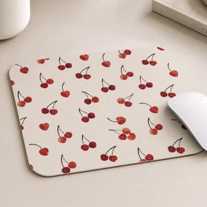 May include: A rectangular mouse pad with a light beige background featuring a repeating pattern of watercolor-style red cherries with dark brown stems. The surface is smooth and flat, displaying multiple pairs of cherries scattered across the design.