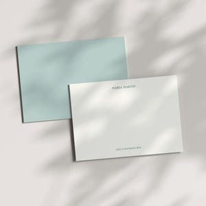 May include: Two stationery cards. One is white with a light blue border and the text "ISABELLA BROWN" and address. The other card is light blue with the initials "IB". The cards are on a white surface with shadows.