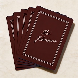 May include: A stack of burgundy coasters with a white border and the text "The Johnsons" in a cursive font. The coasters are arranged in a fan shape, showcasing their design.