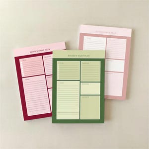 May include: Three daily plan notepads in shades of pink and green. Each notepad has sections for to-do lists, priorities, notes, reminders, and tomorrow. The notepads are labeled "Menia's Daily Plan", "Nicole's Daily Plan", and "Olivia's Daily Plan".