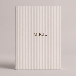May include: A hardcover book featuring a vertical striped pattern in off-white and light beige. The center of the front cover displays the letters M.K.L. in a gold-colored serif font. The book has a clean, rectangular shape with a bound spine.